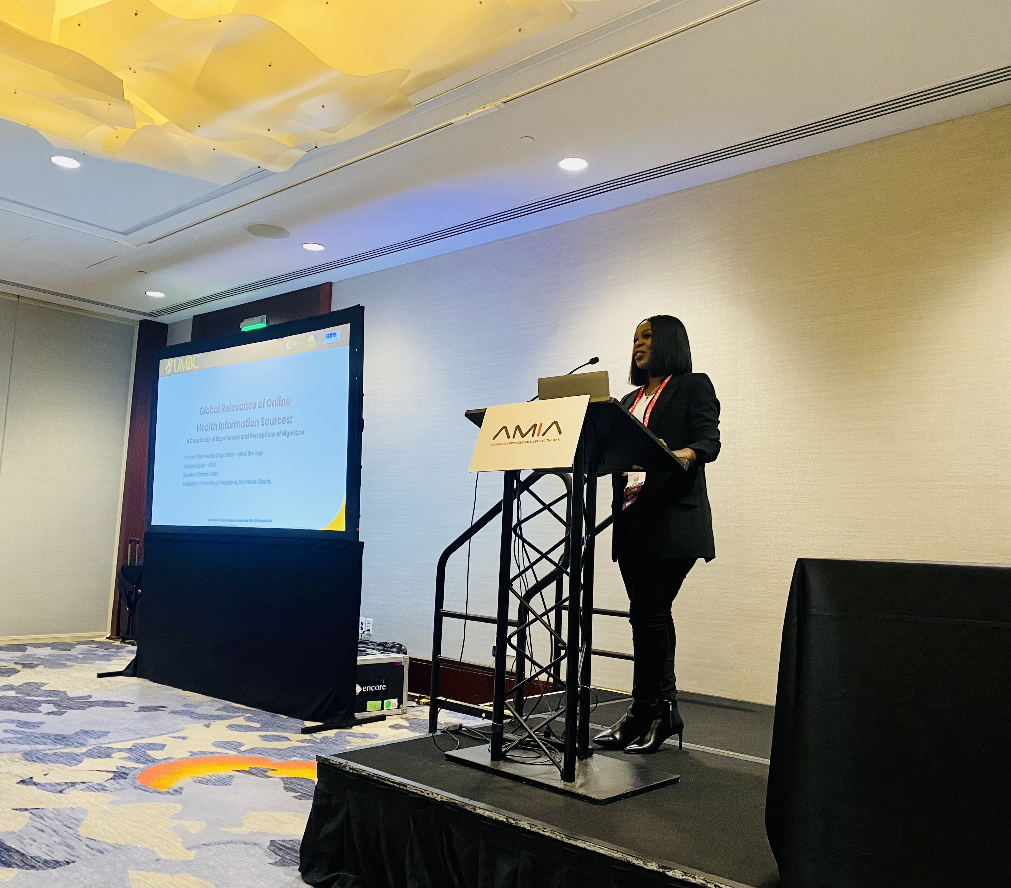 Ommo Clark presenting her research paper at the lectern of the AMIA 2024 Annual Symposium in San Francisco, with a UMBC slide visible on the projection screen